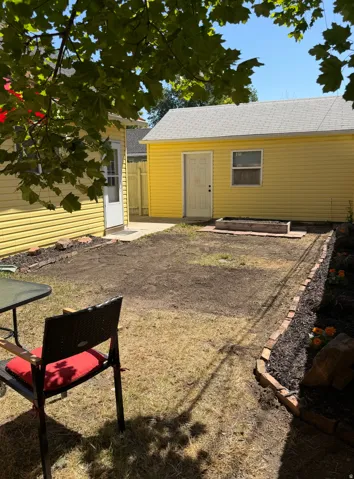 Rear property area featuring a detached yellow siding structure with a gray shingle roof, a concrete pad, and a planting bed