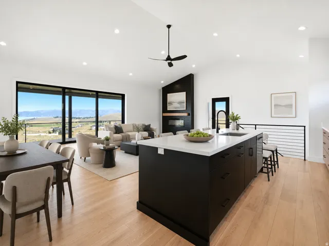 Spacious open-concept living area featuring a large kitchen island with integrated sink, wood-finish flooring, a contemporary ceiling fan, and large sliding glass doors opening to an outdoor railing