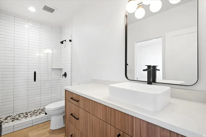 Modern bathroom featuring a wood-finish vanity with a white vessel sink, a large framed mirror, and a walk-in shower with horizontal white subway tiling and a pebble-tiled floor