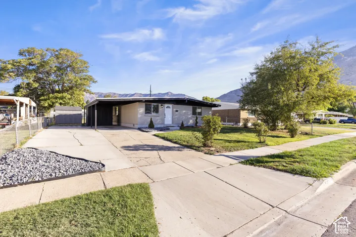 Single-level residence with a painted brick exterior, attached carport, concrete driveway, and front lawn, set against a mountain backdrop