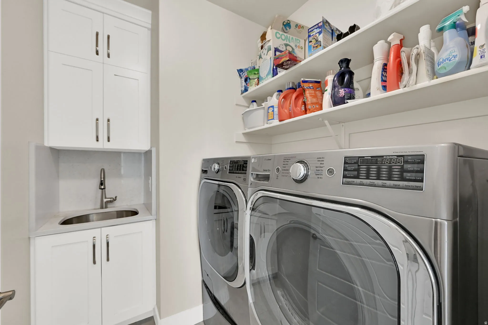 Dedicated laundry area featuring white shaker cabinetry, an undermount sink with a gooseneck faucet, light-toned countertops, and open shelving