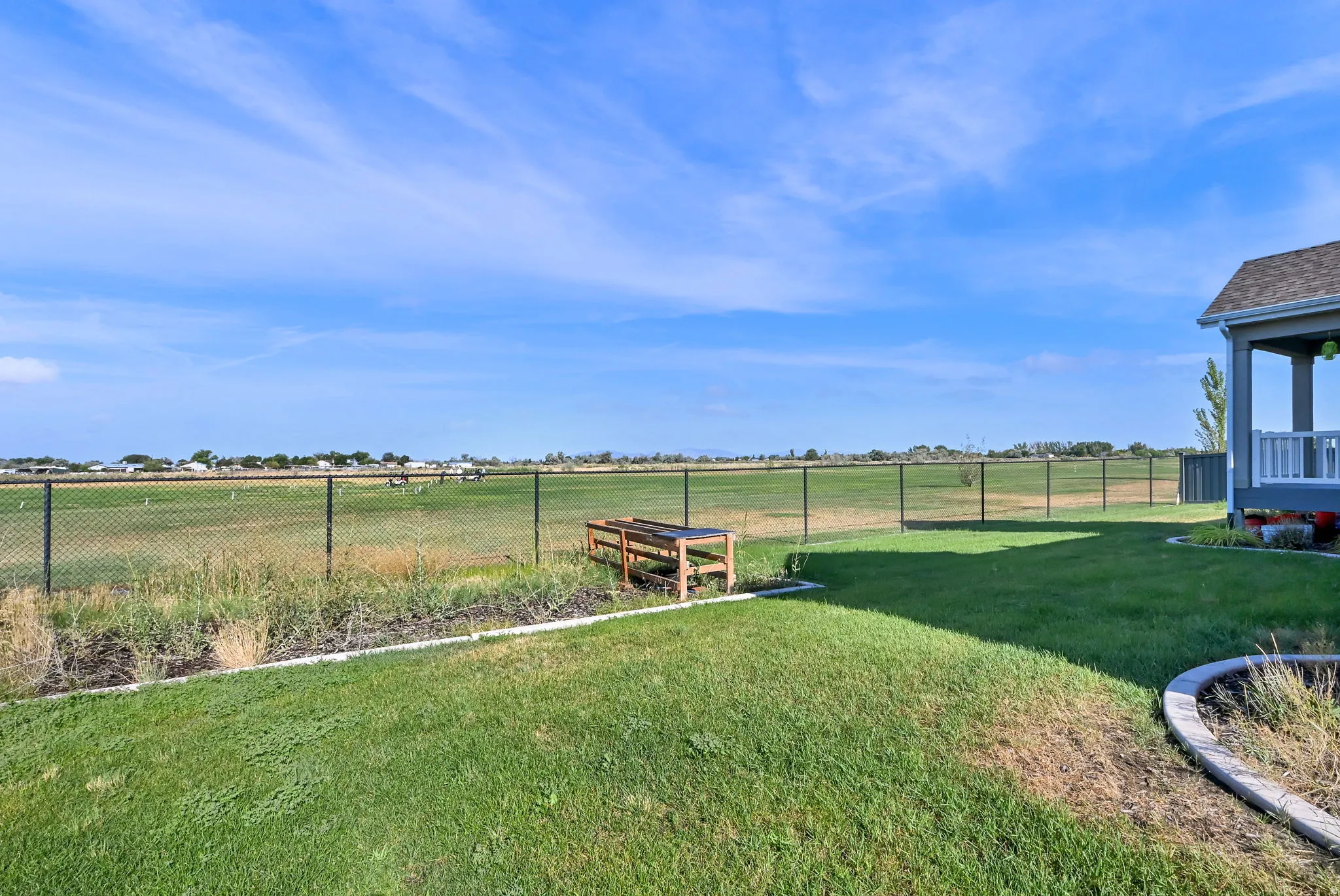 Expansive backyard featuring a green lawn, mature landscaping, a covered patio with white railing, and a black chain link fence