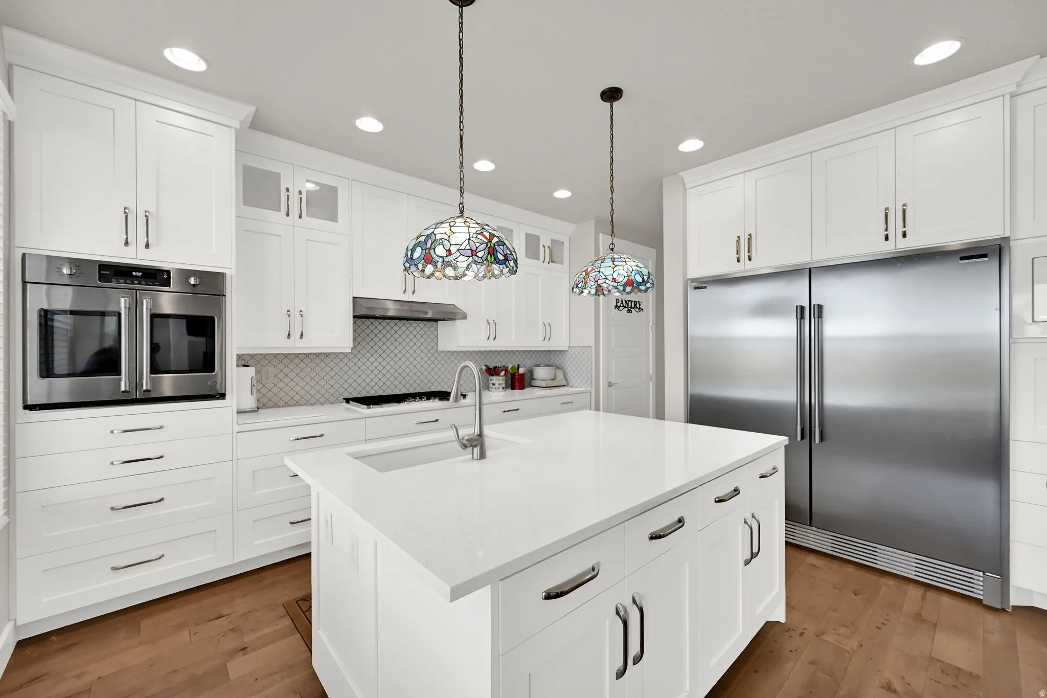 Kitchen featuring a central island with an integrated sink, white shaker cabinetry with brushed nickel hardware, light-toned countertops, and wood-finish flooring