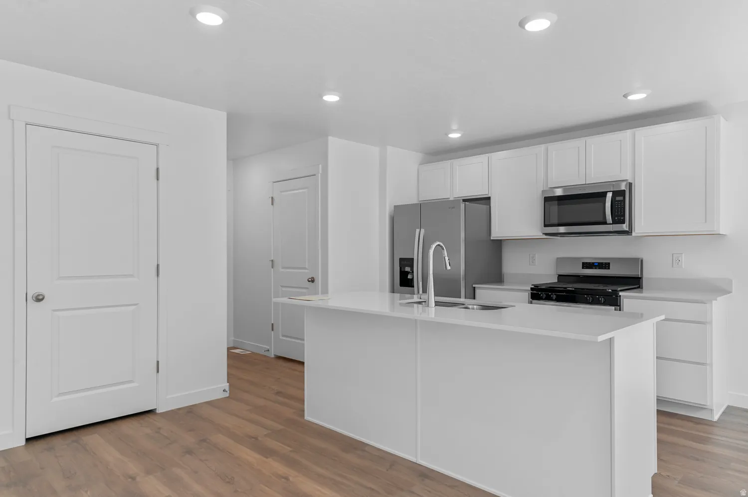Kitchen featuring white cabinetry, stainless steel appliances, a center island with sink, light countertops, and wood-finish flooring
