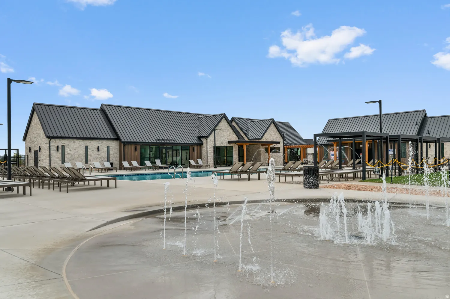 Resort-style pool area featuring a splash pad, multiple lounge chairs, and a contemporary building with a standing seam metal roof and light-toned brick facade