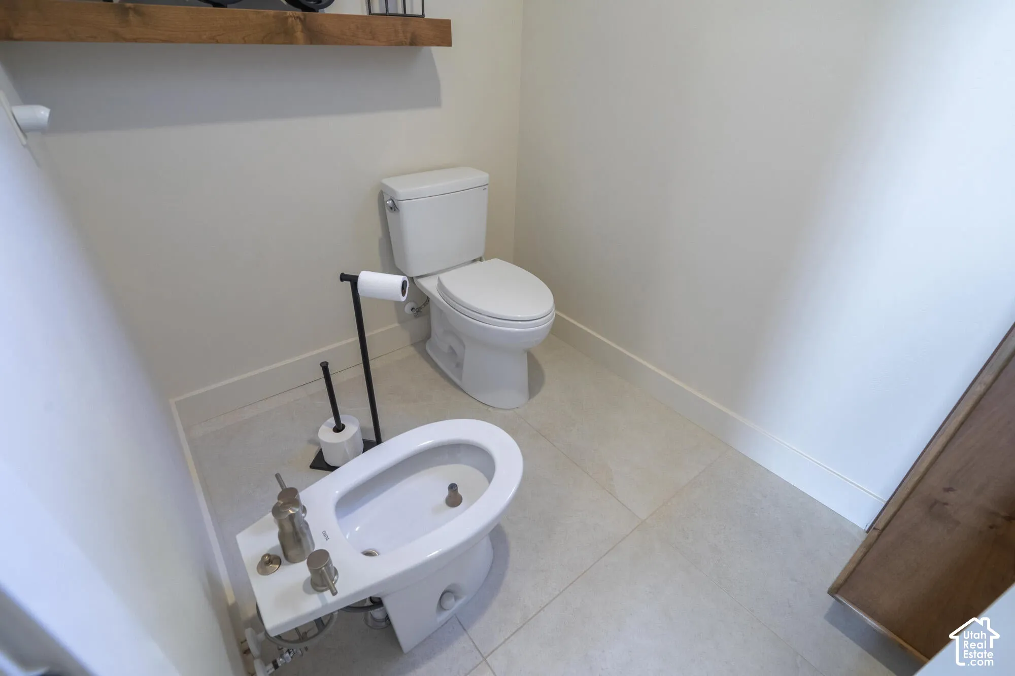 Powder room featuring a bidet, white toilet, light-colored floor tiling, a floating wood-finish shelf, and white walls