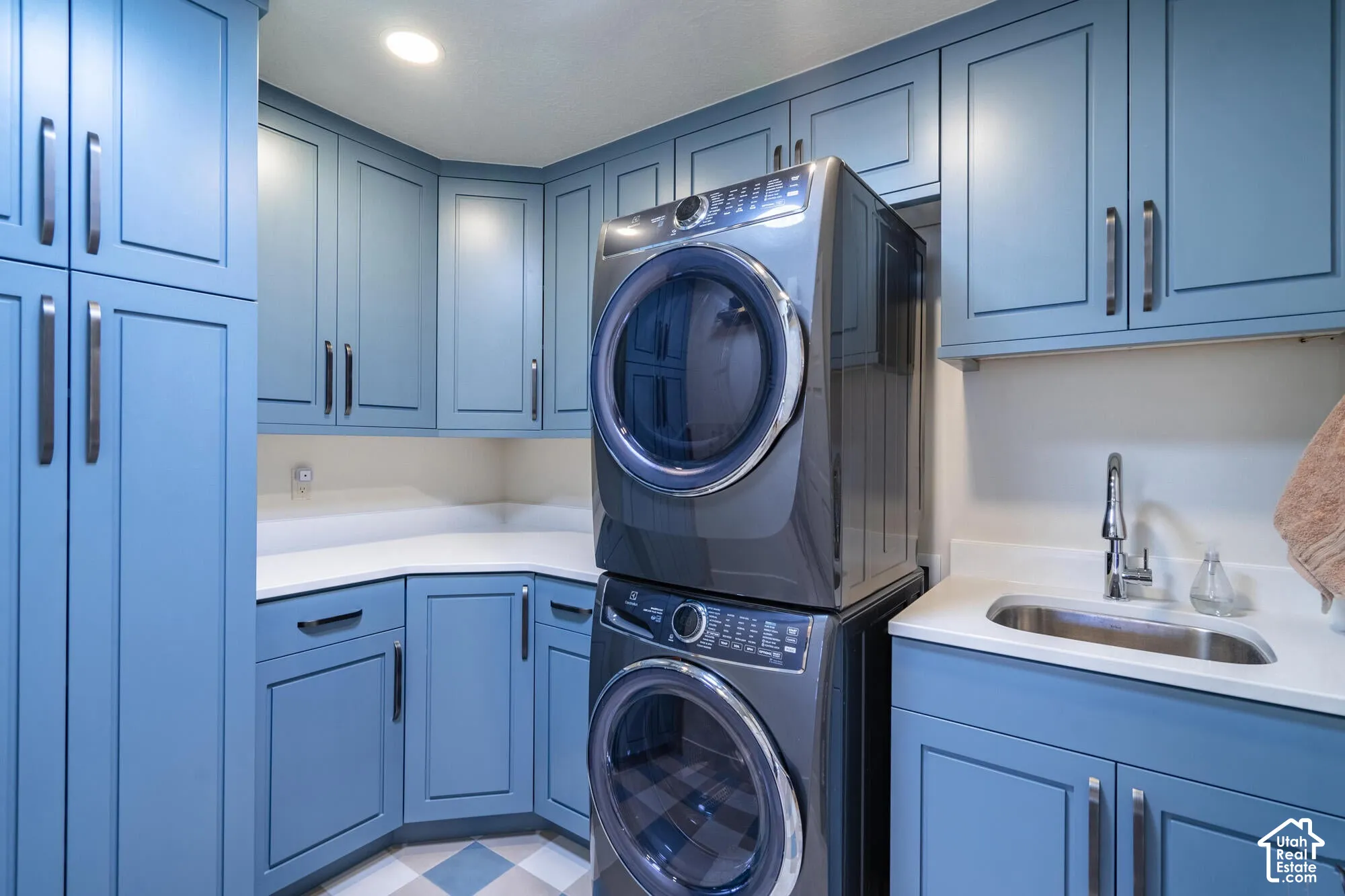Laundry area featuring custom blue cabinetry, light-toned countertops, a stainless steel sink with a gooseneck faucet, and a stacked washer and dryer