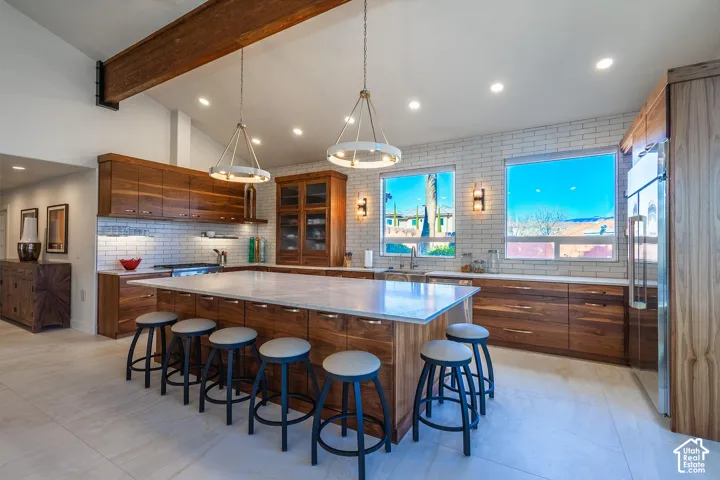 Spacious kitchen featuring a large central island with seating, extensive wood-finish cabinetry, white subway tile backsplash, recessed lighting, and a prominent wood ceiling beam