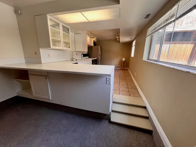 East Side - Kitchen area featuring white cabinetry with glass-front uppers, a stainless steel sink with a gooseneck faucet, and white countertops extending into a peninsula with integrated shelving