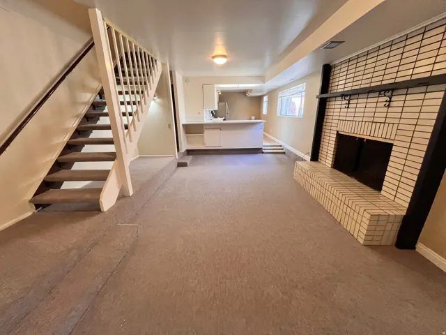 East Side - Spacious room featuring a white brick fireplace with a black mantel, an open staircase with a wooden handrail, and light brown carpeting