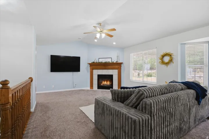 Living area featuring vaulted ceilings, a built-in fireplace with a wood mantel, carpet flooring, a ceiling fan with integrated lighting, and multiple windows with blinds