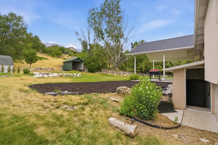 Spacious backyard featuring a covered patio, an additional storage shed, a manicured lawn area, and tiered landscaping with natural rock accents