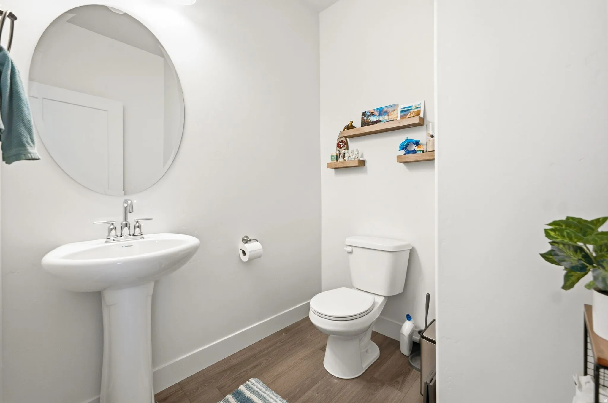 Pedestal sink with chrome faucet, circular wall mirror, wood-finish flooring, and light-toned walls