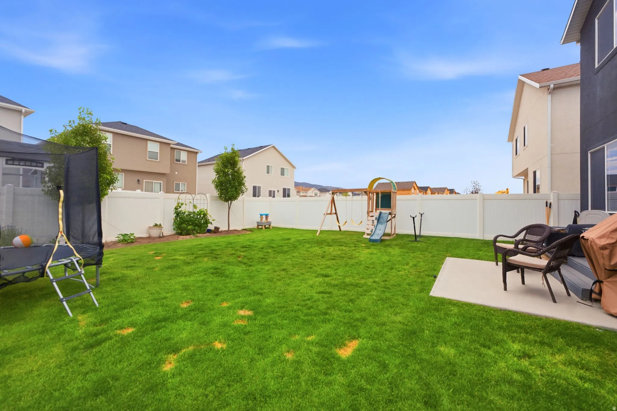 Expansive green lawn with a concrete patio, mature trees, and a white vinyl privacy fence