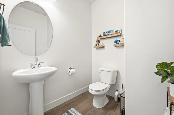 Pedestal sink with chrome faucet, circular wall mirror, wood-finish flooring, and light-toned walls
