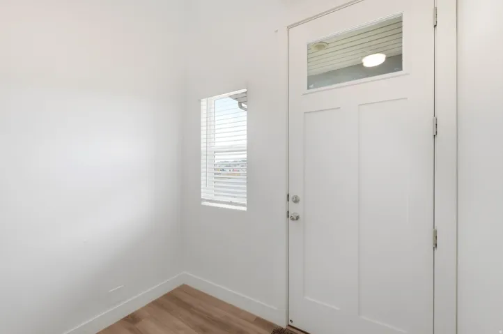 Entryway featuring a white paneled door with an upper glass insert, wood-finish flooring, white baseboards, and a window with horizontal blinds
