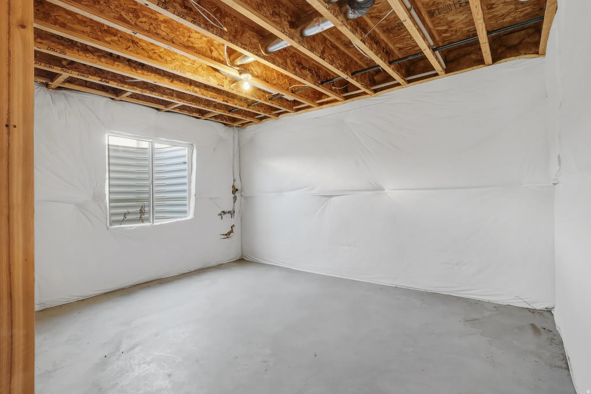 Unfinished basement featuring exposed wood joists, concrete flooring, insulated walls, and a window