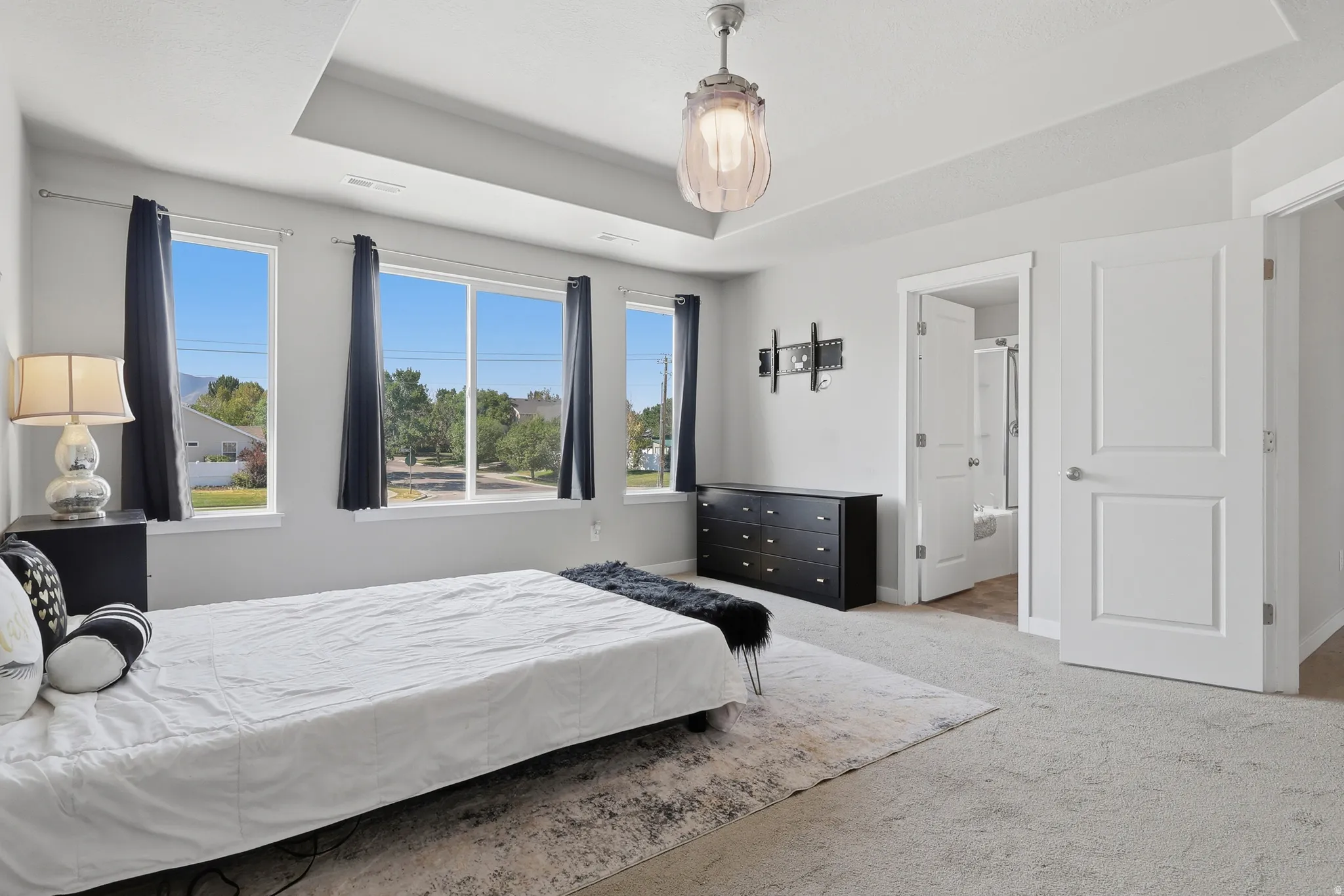 Spacious interior room featuring light gray carpeting, three large windows, a tray ceiling, and a contemporary ceiling fixture
