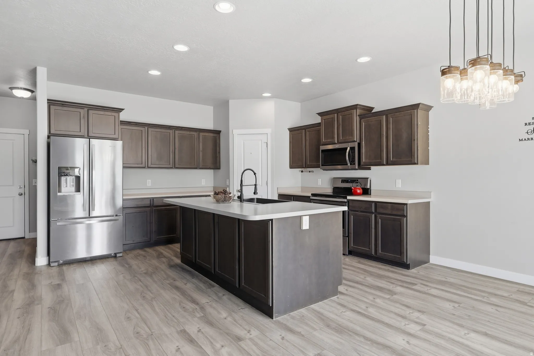 Kitchen featuring wood-finish flooring, dark wood cabinetry, white countertops, a stainless steel refrigerator, and a kitchen island with an integrated sink