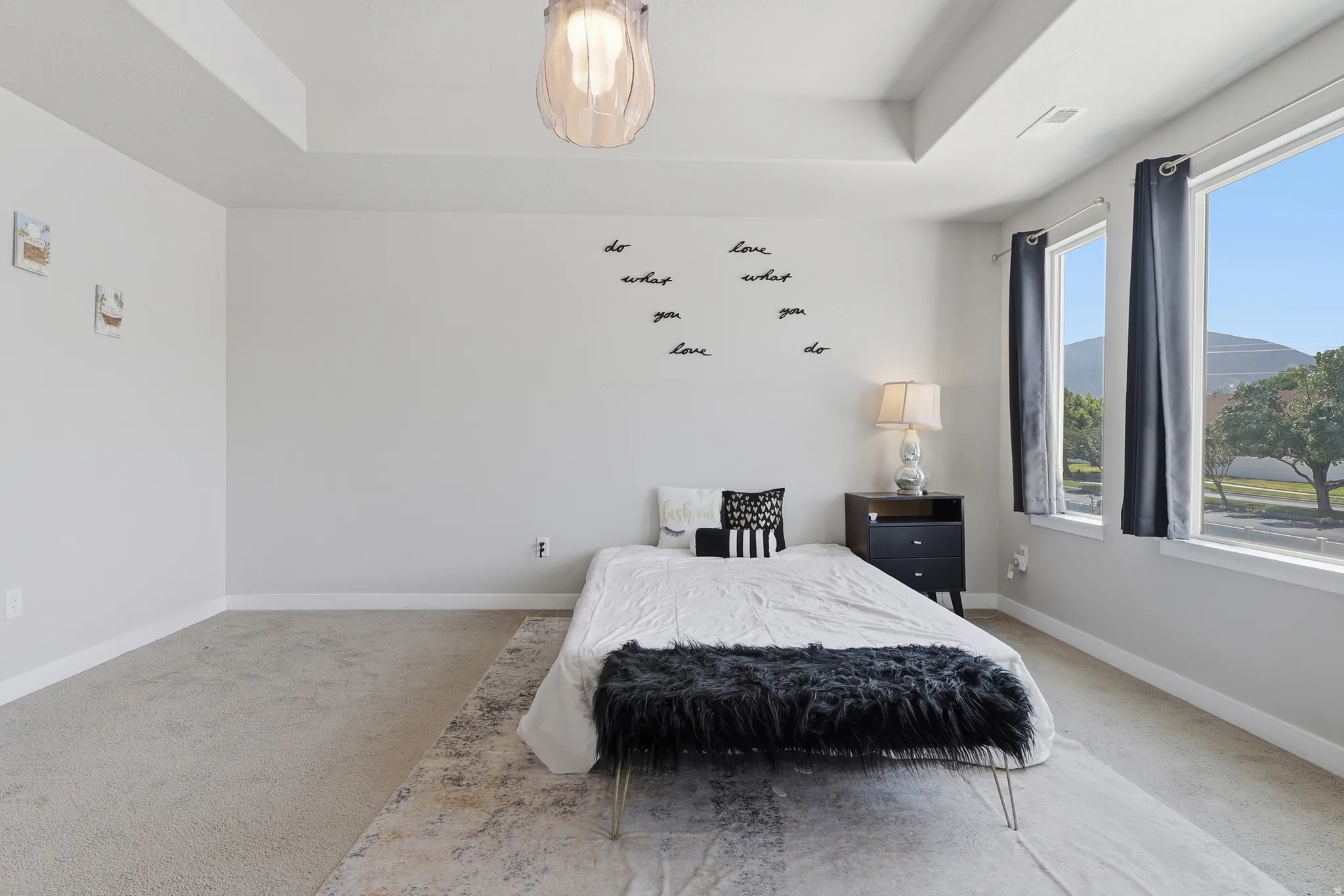 Spacious bedroom featuring light neutral wall paint, tray ceiling, two large windows, and light-colored carpet flooring
