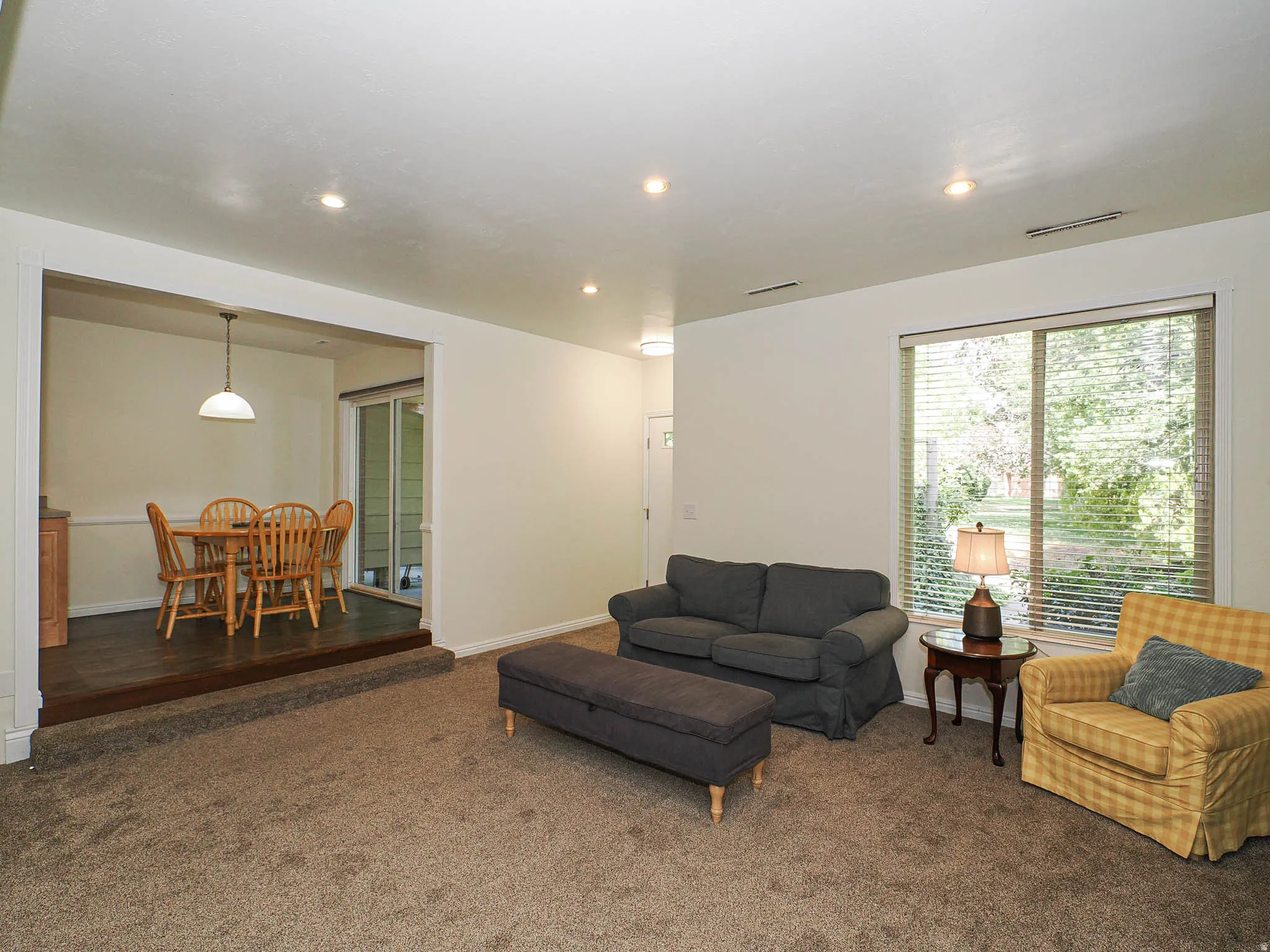 Carpeted living area featuring recessed lighting, a large window with blinds, and a built-in step leading to a separate dining area with wood-finish flooring and a sliding glass door