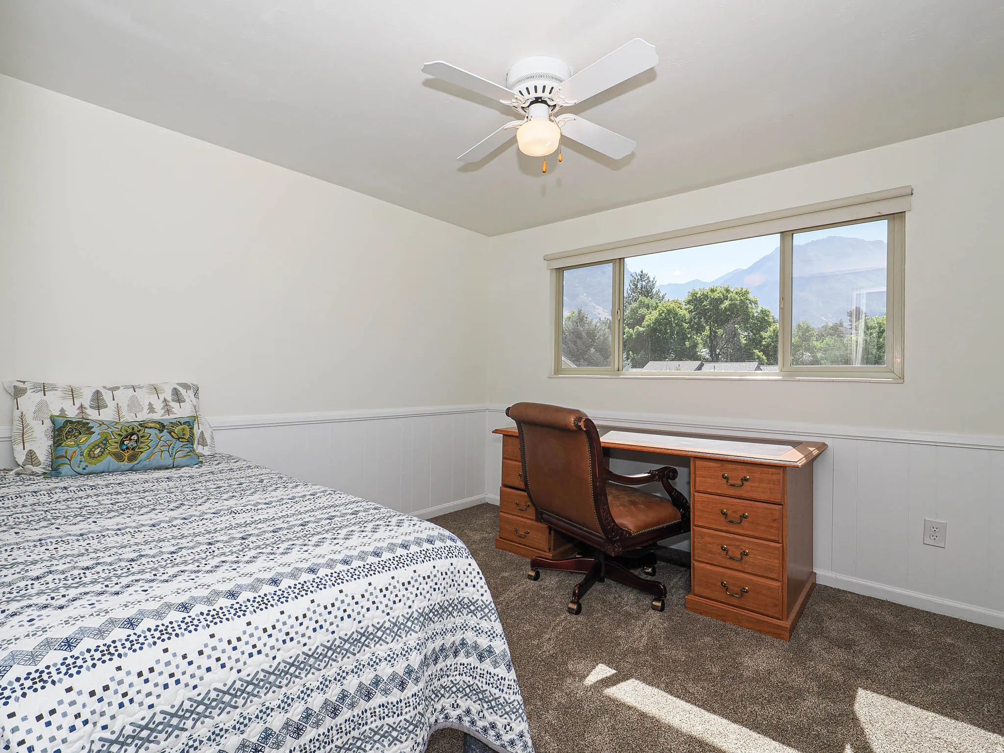 Window offering mountain scenery, white wall paneling, ceiling fan with light fixture, light neutral wall paint, and textured carpet flooring