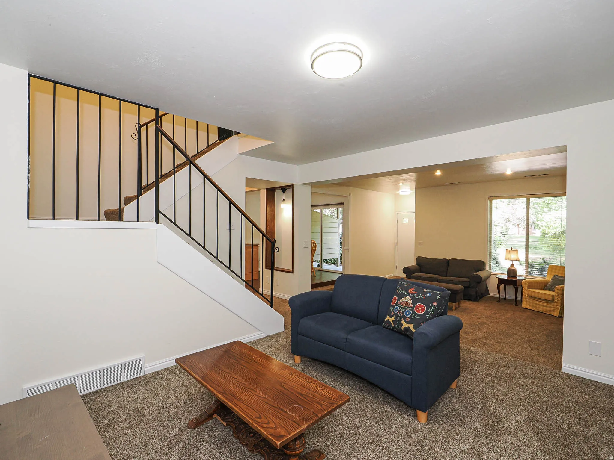 Open-concept living area featuring neutral-toned walls, plush carpeting, and a built-in staircase with wrought iron railing