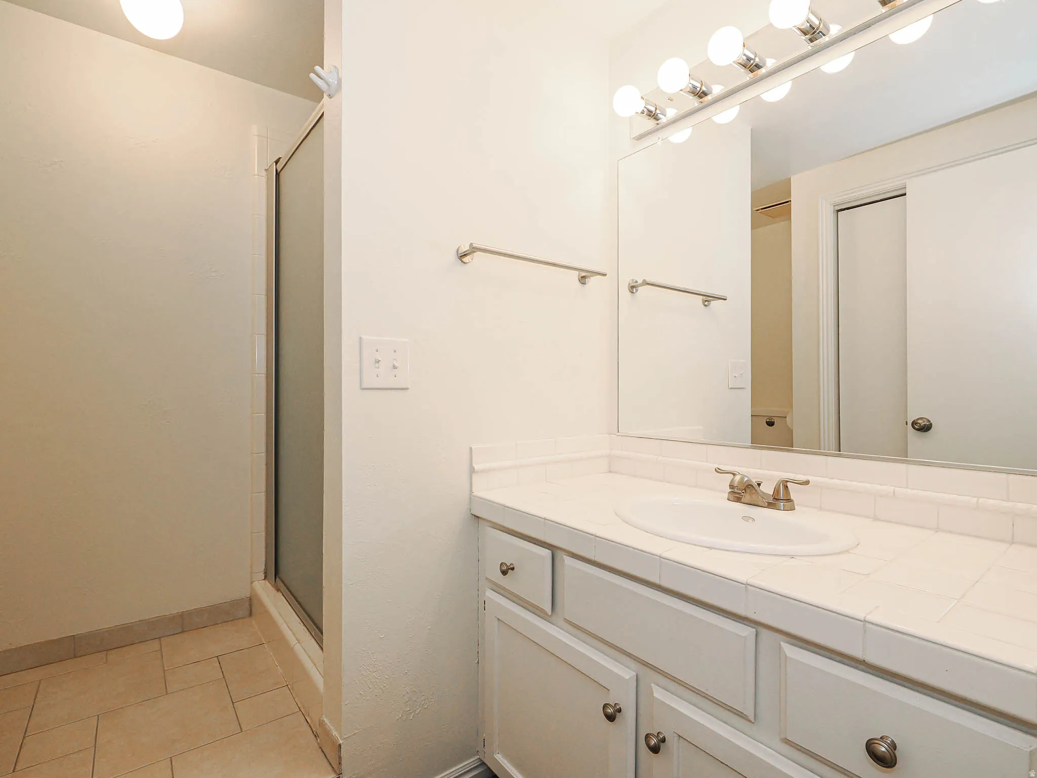Bathroom featuring a tile-top vanity with an integrated sink and brushed nickel faucet, a large mirror with strip lighting, a glass-enclosed shower, and tiled flooring