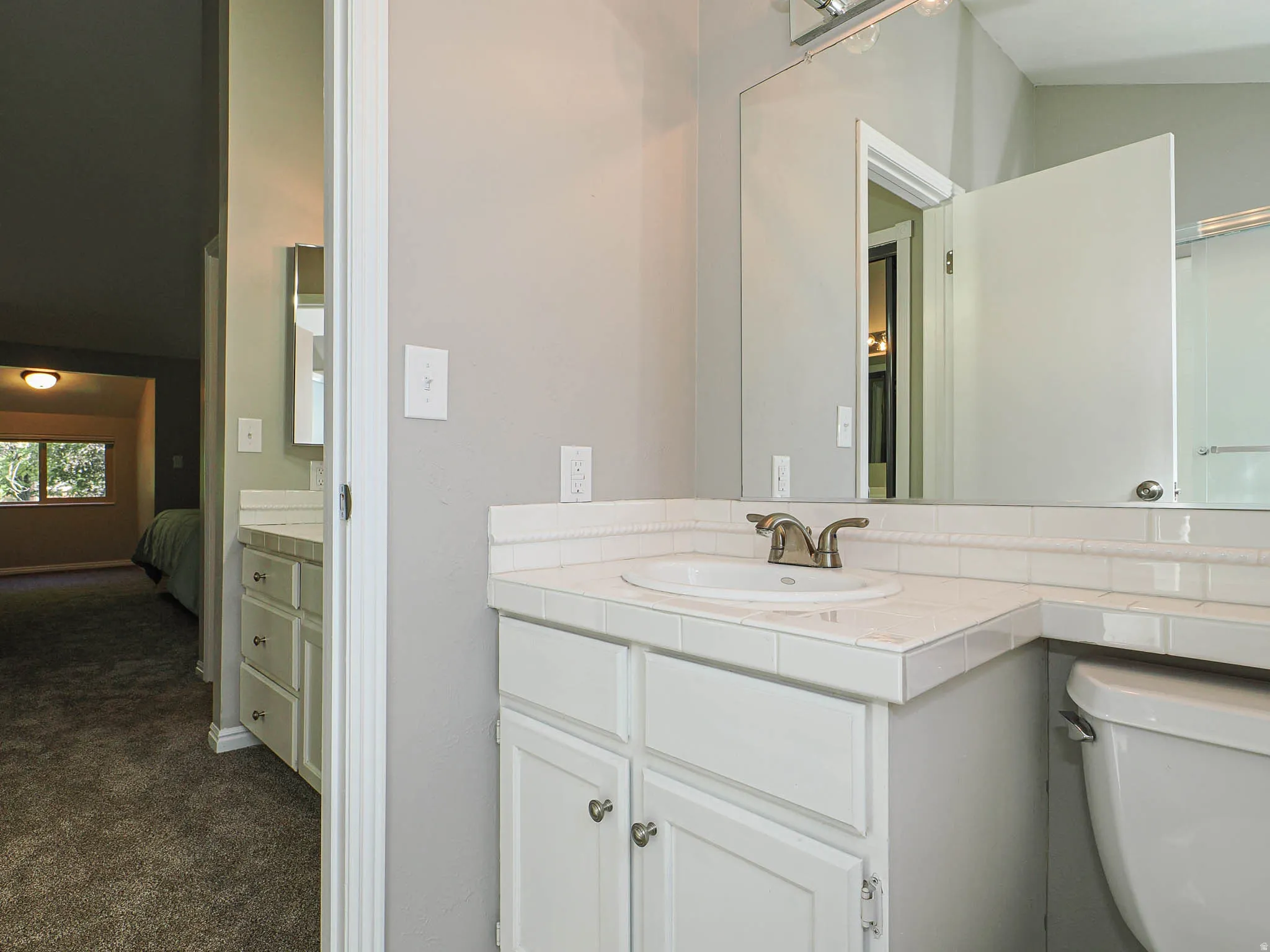 Bathroom vanity with white tile countertop and integrated sink, bronze-finish faucet, and white paneled cabinetry