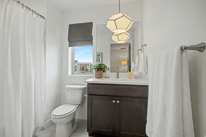 Bathroom featuring a modern geometric pendant light, a dark wood-finish vanity with a white countertop and undermount sink, a large wall mirror, and a window with a Roman shade
