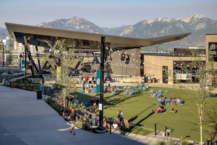 Expansive outdoor gathering space featuring a modern stage structure, manicured green lawn, string lighting, and mountain backdrop
