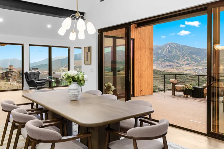 Open-concept dining space with a contemporary chandelier, multiple floor-to-ceiling windows, light wood-finish flooring, and direct access to an outdoor deck featuring panoramic mountain scenery