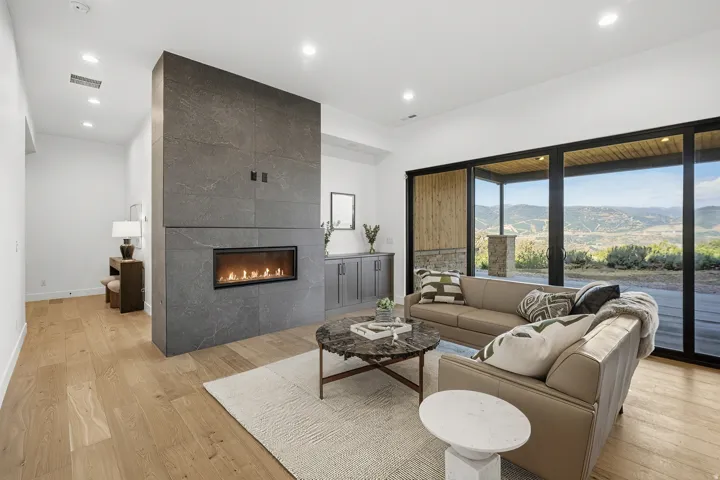 Contemporary living area featuring a linear fireplace set within a floor-to-ceiling stone-finish wall, expansive sliding glass doors, light wood-finish flooring, recessed lighting, and a built-in cabinetry with a countertop
