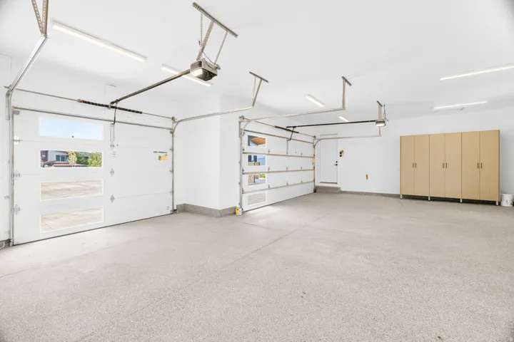 Spacious garage featuring a light gray epoxy floor coating, multiple white garage doors with upper window panels, integrated ceiling lighting, and built-in wood-finish cabinetry