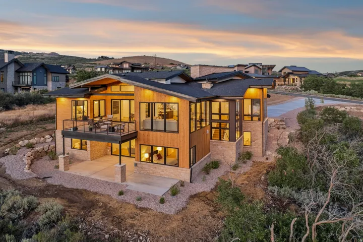 Modern architectural design featuring extensive window walls, a spacious upper-level balcony, a lower-level patio, natural wood-finish siding, and stone accents
