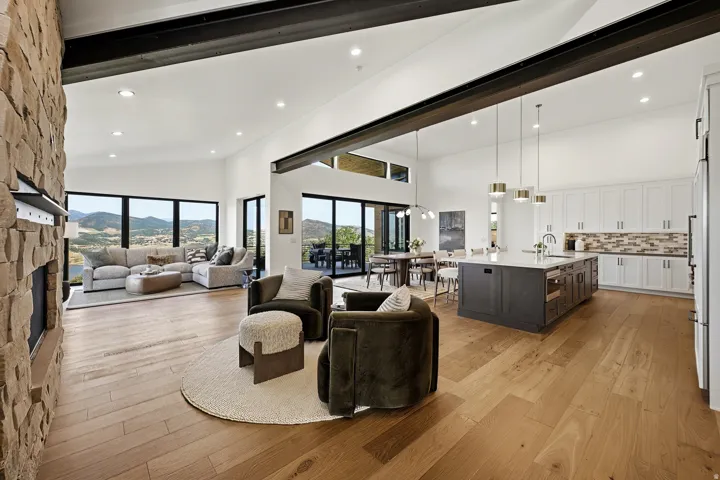 Open-concept living space featuring high vaulted ceilings with exposed structural beams, wood-finish flooring, a stone-clad fireplace, a kitchen island with seating, and a wall of expansive windows
