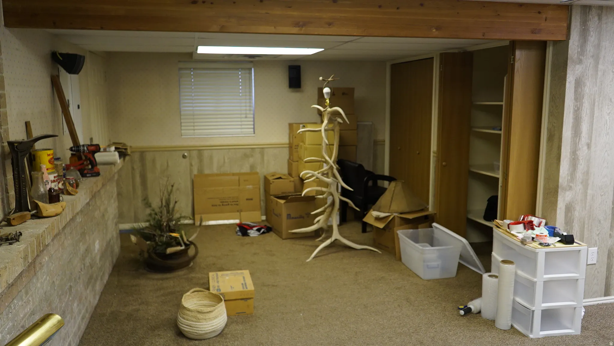 Carpeted room featuring a drop ceiling with recessed lighting, a window with horizontal blinds, built-in cabinetry with shelving, a wood-finish overhead beam, and a brick half-wall