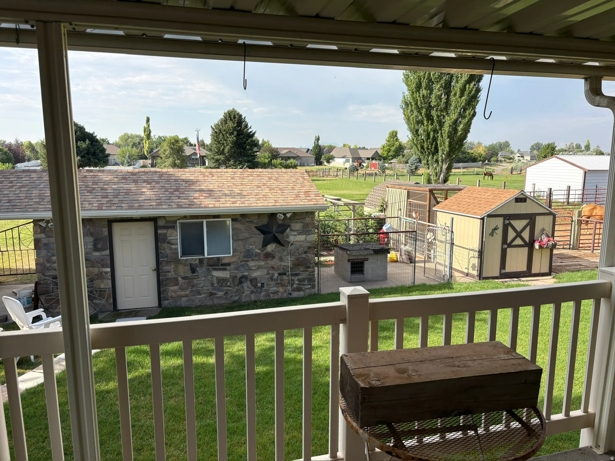 Covered patio featuring white railings, overlooking a stone-clad outbuilding with a shingle roof, two storage sheds, and fenced pasture areas