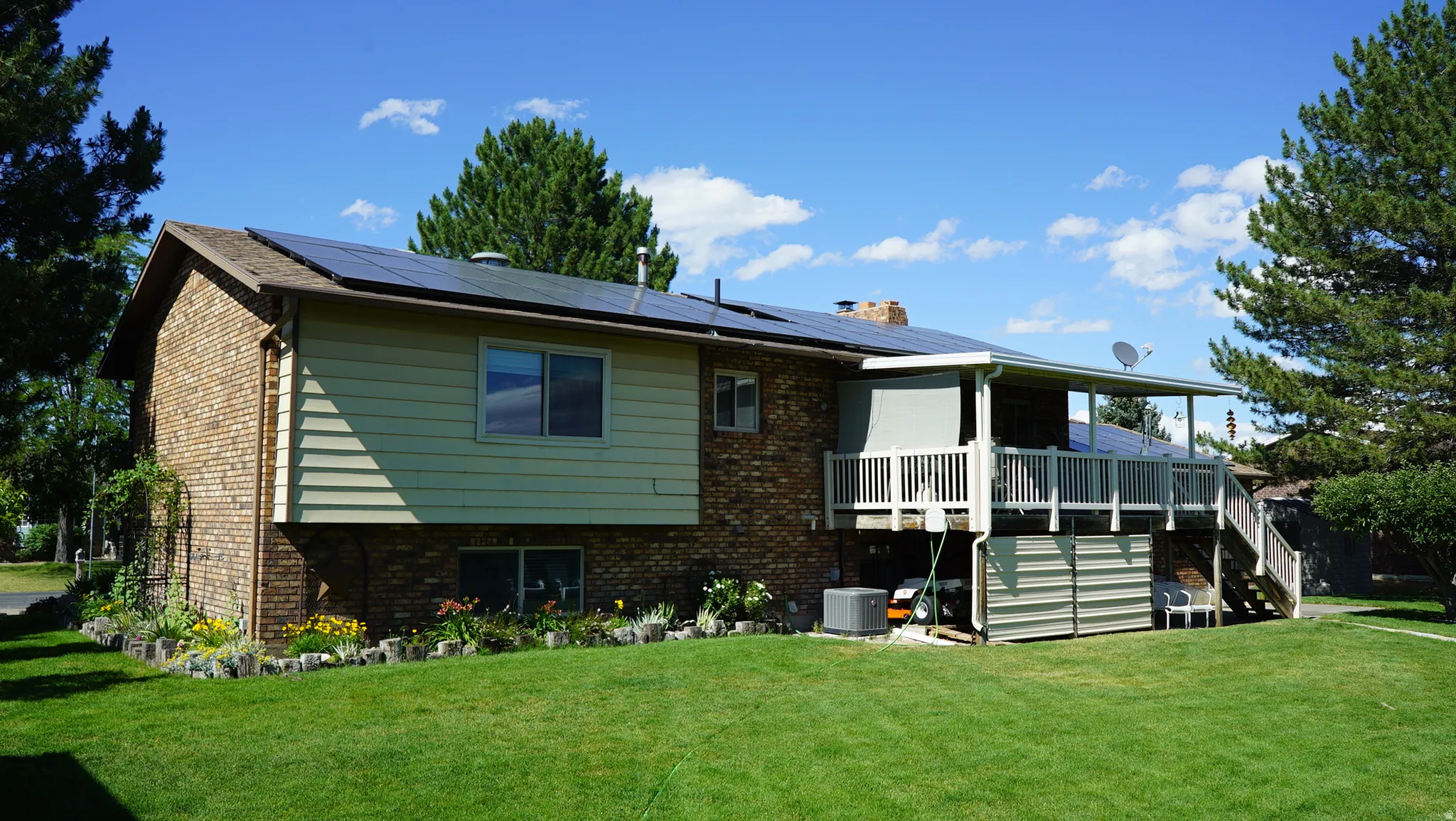 Rear exterior featuring a brick and siding facade, a covered upper-level deck with white railings, solar panels on the roof, a manicured lawn, and mature trees
