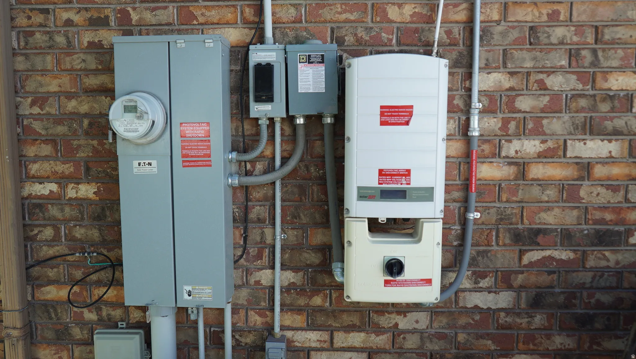 Exterior brick wall featuring an electrical meter, multiple grey electrical boxes, and a white solar inverter unit