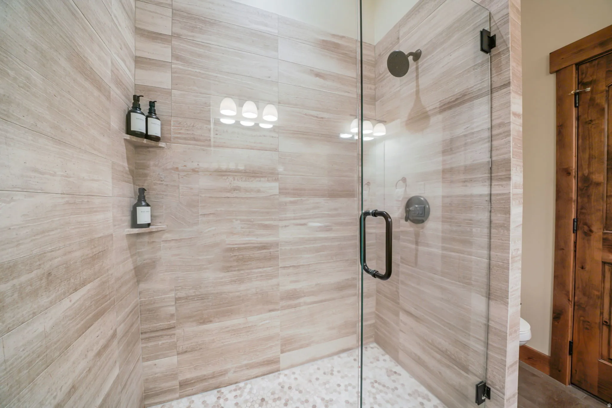 Walk-in shower featuring glass enclosure, stone-look tile walls, pebble-finish shower pan, and built-in shelving