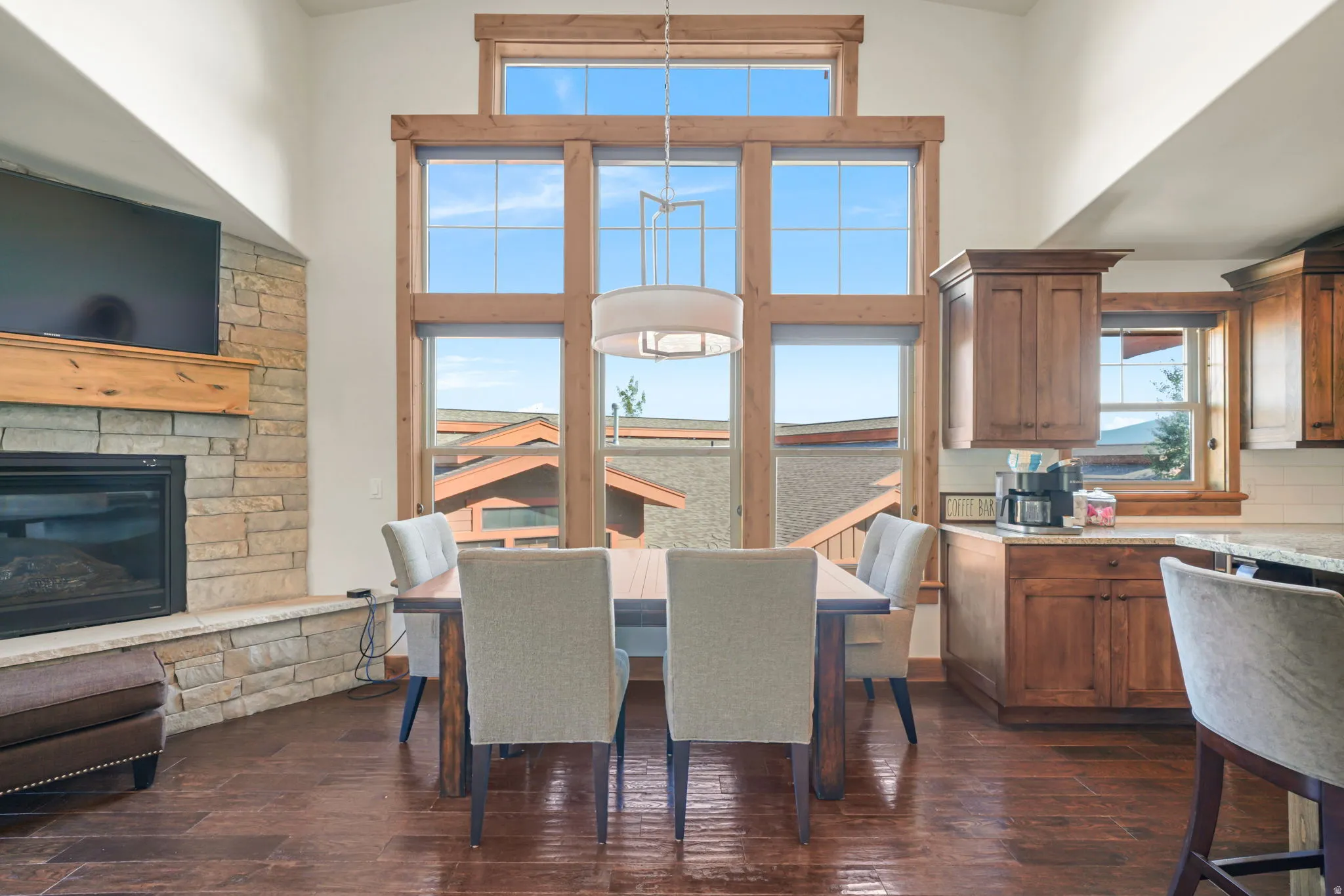 Vaulted ceilings enhance a living area featuring a stacked stone fireplace with a wood mantle and a large window wall with natural wood trim