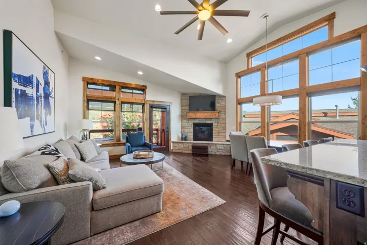 Vaulted ceiling great room featuring a stone fireplace with wood mantel, a wall of windows, dark wood-finish flooring, and a kitchen island with a light-toned countertop