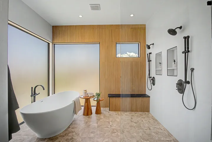 Freestanding soaking tub with a floor-mounted faucet, dual shower systems with matte black fixtures, integrated shower niches, a built-in shower bench, and a wood-finish accent wall