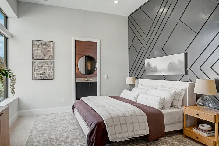 Spacious bedroom featuring a dramatic accent wall with geometric paneling, an en-suite bathroom with a vanity and round mirror, a large window with transom, and recessed lighting