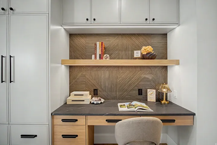 Built-in desk area featuring a dark countertop, light wood cabinetry with black hardware, and a geometric wood-finish accent wall