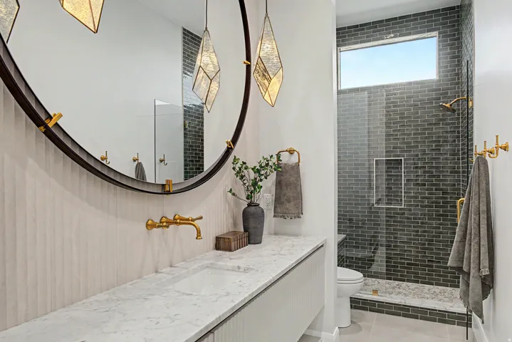 Bathroom featuring a large circular mirror with pendant lighting, a single vanity with a quartz countertop, a wall-mounted brass faucet, and a glass-enclosed shower with dark subway tile and a horizontal privacy window