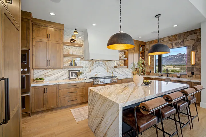 Gourmet kitchen featuring a waterfall island with stone countertop, natural wood cabinetry, built-in double ovens, a gas range, and a stone accent wall