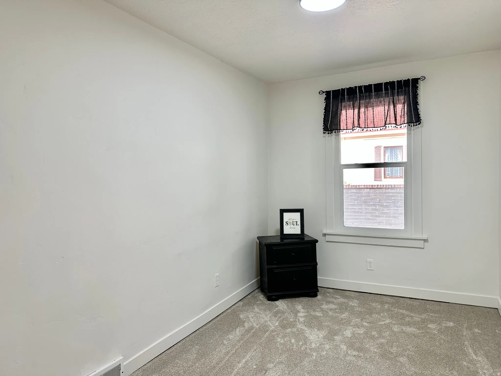Bright room featuring a window with white trim, light-colored carpet flooring, white walls, and a ceiling-mounted light fixture