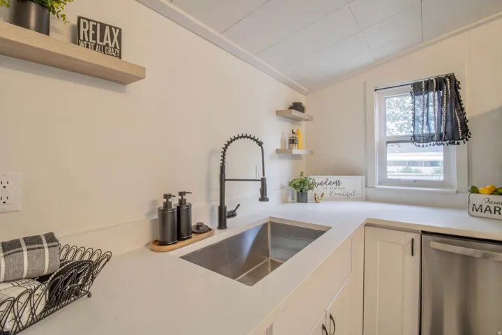 Kitchen featuring a stainless steel, single basing, undermount sink, a matte black gooseneck faucet, light-toned countertops, white cabinetry, and floating shelves